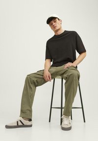Zwart T-shirt met korte mouwen, olijfgroene cargo-broek en beige sneakers met zwarte accenten. Model zit op een groene kruk.