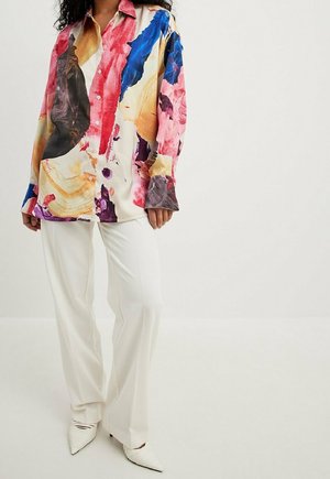 Kobieta ubrana w białe spodnie, białe szpiczaste buty oraz luźną, wielokolorową koszulę z abstrakcyjnym nadrukiem i guzikami.