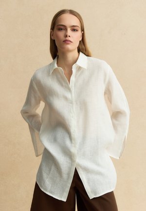 Mujer con pelo rubio liso lleva una camisa blanca de lino de gran tamaño abotonada y pantalones marrones, de pie contra un fondo beige.