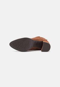 Botte de cheville en suede marron avec une semelle texturée, un talon effilé et un intérieur lisse. Le design est doté d'une fermeture éclair sur le côté.
