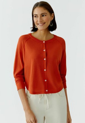Femme souriante, portant un cardigan orange boutonné à manches trois-quarts et un pantalon beige clair avec cordon de serrage, fond uni clair.