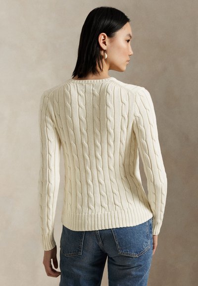 Femme aux cheveux noirs et raides portant un pull en maille torsadée crème et un jean bleu, vue de profil sur un fond beige uni.