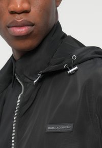 Μαύρο ελαφρύ μπουφάν με κουκούλα, με ασημένιο φερμουάρ, διακοσμητικά κορδόνια και επωνυμία στο στήθος. Λεία υφή.