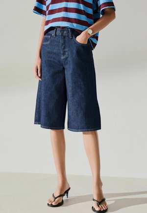 Person wearing dark blue wide-leg denim shorts, striped short-sleeve shirt, wristwatch, and black heeled sandals, standing with one hand in pocket.