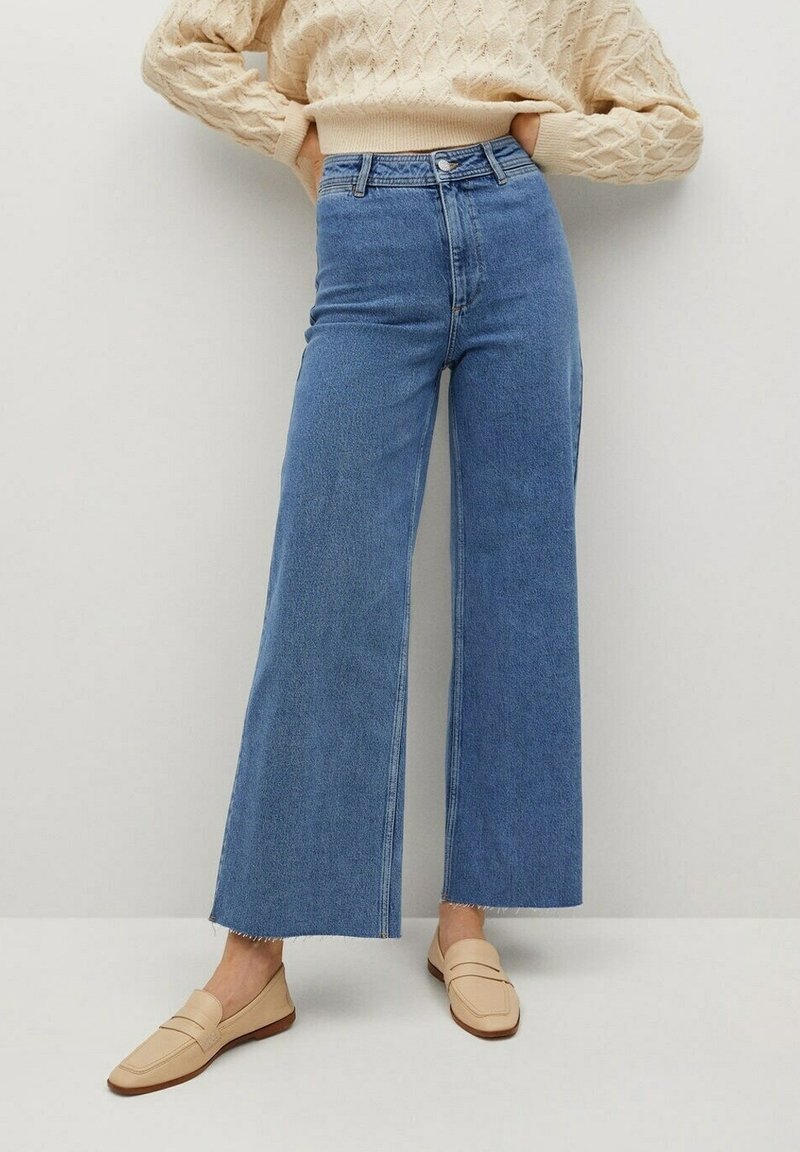 Personne portant un jean bleu taille haute à jambes larges avec des ourlets bruts, des mocassins beige et un pull en maille torsadée crème, debout contre un mur clair uni.