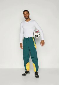Groene en gele trainingsbroek met een trekkoord in de taille, voorzien van zijaccenten. Witte langemouwen shirt. Voetbal aan de zijkant gehouden. Zwarte schoenen.