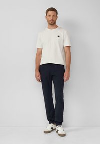 Witte katoenen T-shirt met een klein zwart logo, gecombineerd met marineblauwe broeken en witte sneakers met bruine accenten en een gestructureerde zool.