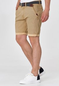 Shorts beiges en coton avec une texture rayée, des revers roulés et une ceinture sombre à motifs. Associés à des chaussures blanches avec des accents noirs.