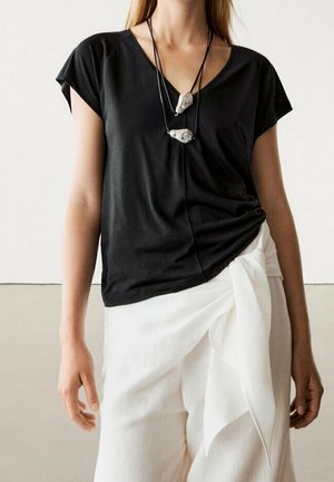 Frau trägt ein schwarzes V-Ausschnitt-Shirt, in weiße hoch taillierte Hose gesteckt, mit einem Knoten und zwei Steinanhänger-Halsketten an schwarzen Bändern.