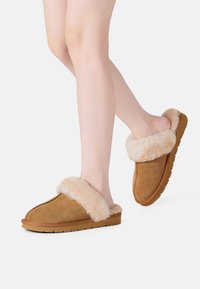 Tan suede slippers with plush cream fur cuffs, flat sole, rounded toe, and visible stitching detail. Lightweight and soft texture.