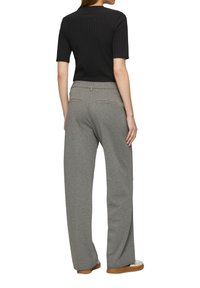Femme de dos portant un haut noir à manches courtes, un pantalon large gris et des chaussures blanches avec des semelles beiges sur fond blanc.