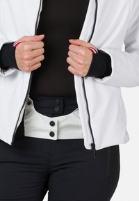 Giacca softshell bianca, aperta, abbinata a pantaloni neri. La giacca presenta polsini neri con strisce rosse e bianche, evidenziando una texture del tessuto liscia.