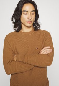 Maglione a coste marrone con scollatura rotonda e maniche lunghe. Presenta un piccolo logo sul petto. La trama è tessuta e elastica.