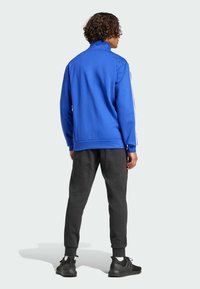 Blå zip-up jacka med hög krage, vita ränder på ärmarna, kombinerad med grå sweatpants och svarta sneakers mot en ljus bakgrund.