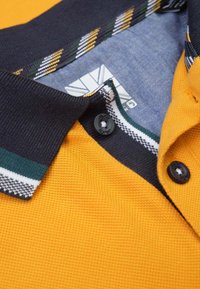 Geel polo-shirt met een gestructureerde stof, marineblauwe kraag met groene en gele strepen, en twee zwarte knopen bij de hals.