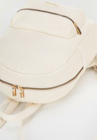 Mochila blanca de tejido trenzado con forma redondeada, dos compartimentos con cremallera y herrajes dorados, y correas ajustables. Superficie suave y texturizada.
