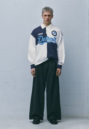 Mladý muž stojí u světlé zdi, má na sobě modrobílý rugby dres Detroit Lions a široké černé kalhoty s černými botami.