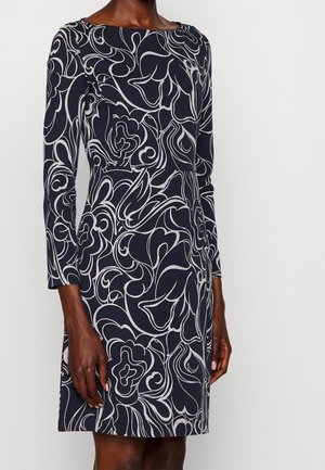 Woman wearing a navy blue long-sleeve dress with a white abstract floral pattern, standing against a plain light background.