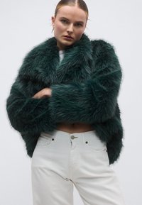 Πράσινο σακάκι από συνθετική γούνα με φαρδιά γιακά και μακριά μανίκια, συνδυασμένο με λευκό τζιν ψηλόμεσο, με κούμπωμα με κουμπιά και τσέπες.