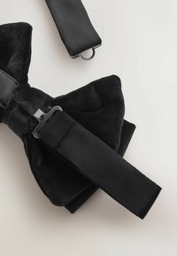 Čierny zamatový motýlik s hladkým saténovým remienkom a kovovou sponou, ktorý má textúrovaný povrch a široký, symetrický dizajn mašle.