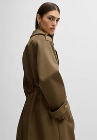 Trench coat marrone con una vestibilità rilassata, vita con cintura e polsini abbottonati. Tessuto morbido e colletto a risvolto; vista laterale del modello.