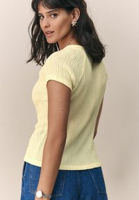 Haut jaune à côtelés avec des manches courtes, doté d'un tissu texturé. Modèle le porte avec un jean en denim taille haute et une montre en argent.