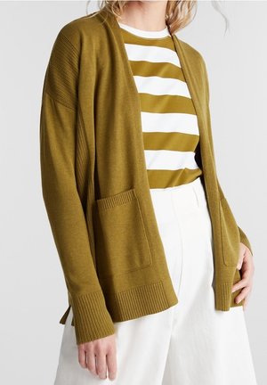 Femme portant un cardigan ouvert vert olive avec poches avant, par-dessus une chemise rayée blanche et vert olive, et un pantalon blanc taille haute.