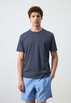 Jonge man staat tegen een effen achtergrond, draagt een donkergrijs T-shirt met korte mouwen en lichtblauwe korte broek, één hand in de zak.
