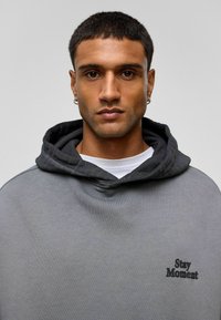 Grijze capuchontrui met een textuurafwerking en een zwart geborduurd "Stay Moment" logo aan de linkerkant, gelaagd over een wit shirt.