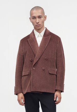 Homme portant un blazer en velours côtelé marron croisé sur une chemise blanche avec un pantalon noir, debout devant un fond clair uni.