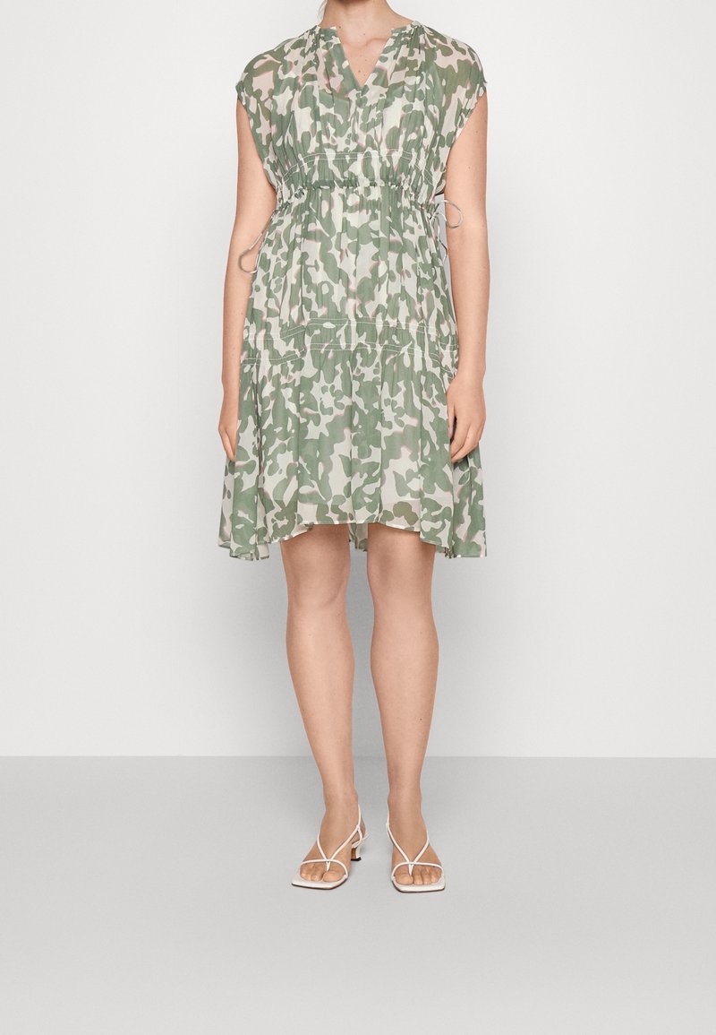 Femme portant une robe ample, longueur genoux, imprimé camouflage vert et beige, manches courtes, et des sandales blanches à talons bas avec brides.