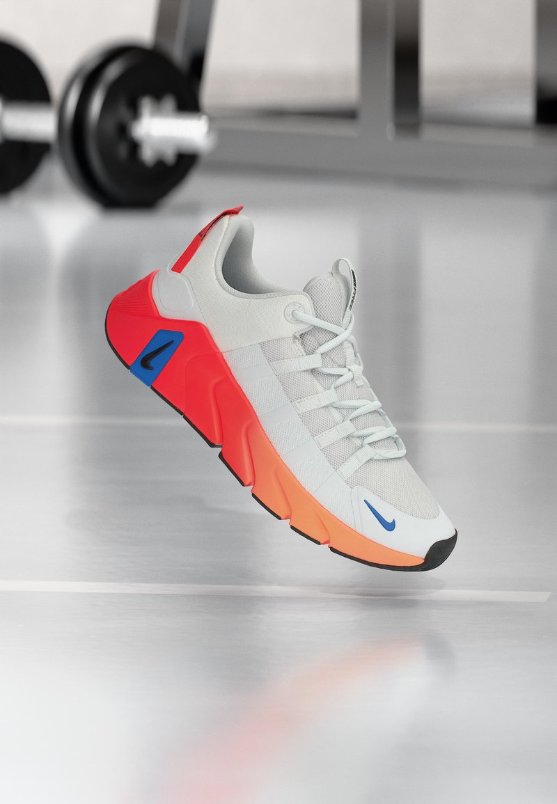 Άσπρο και πορτοκαλί αθλητικό παπούτσι με μπλε λογότυπα Nike, να αιωρείται πάνω από δάπεδο γυμναστηρίου με θολωμένα αλτήρες στο φόντο.