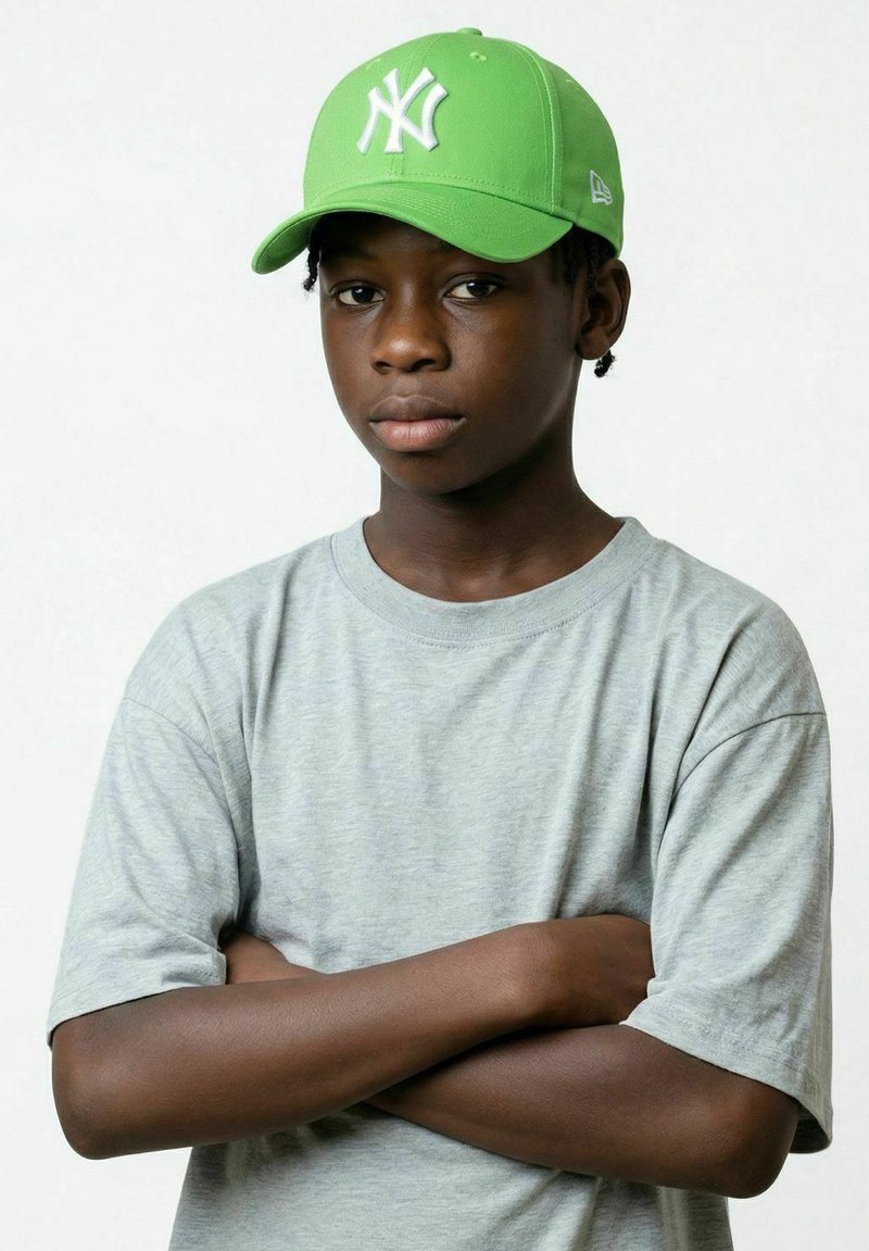 Garçon les bras croisés portant une casquette de baseball vert vif et un t-shirt gris clair, face à un fond uni.