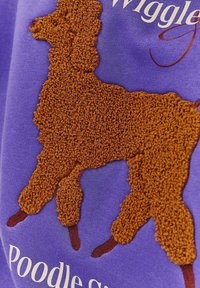 Fialová textilie s texturovaným designem hnědého pudla s nadýchanými smyčkami. Obsahuje vyšívaný text pod tvarem pudla.