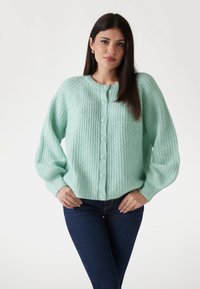 Mintgrüner, strukturierten Strickcardigan mit rundem Hals und sechs Knopfverschluss. Mit Raglanärmeln und gerippten Bündchen. Über dunklen Jeans getragen.