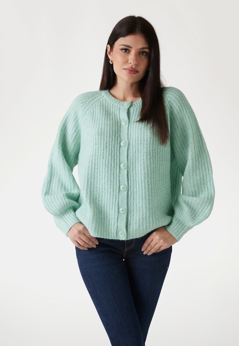 Mintgrüner, strukturierten Strickcardigan mit rundem Hals und sechs Knopfverschluss. Mit Raglanärmeln und gerippten Bündchen. Über dunklen Jeans getragen.