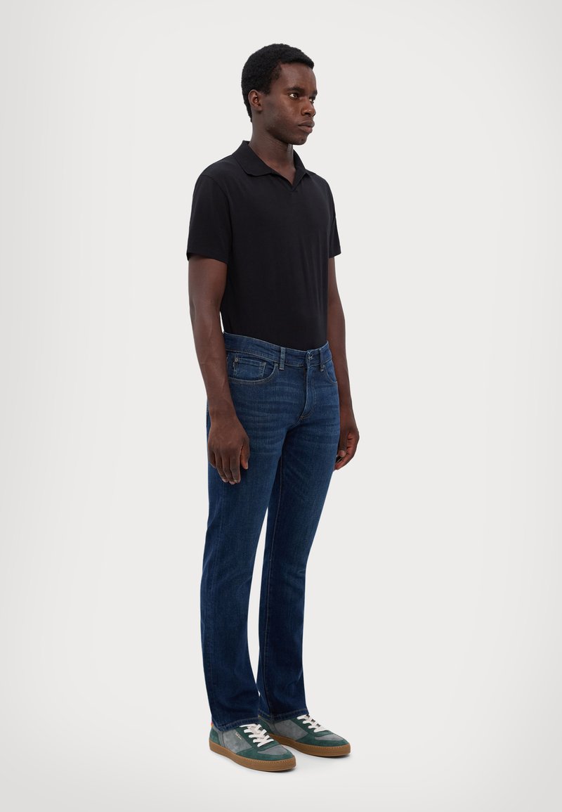 Black short-sleeve polo, blue denim jeans, and multicolored sneakers with white laces. Model stands upright against a light background.