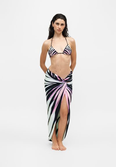 Femme debout pieds nus portant un haut de bikini rayé rose, noir et blanc et un paréo assorti avec une fente à l'avant, sur fond uni.