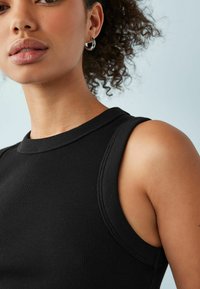 Sort ribbet tanktop med en rund halsudskæring og ærmeløs design, viser et nærbillede af ærmegab og skulderområde. Blød tekstur.