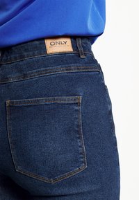 Jeans de mezclilla de color azul oscuro con un bolsillo trasero visible y una etiqueta de cuero marrón cosida en la cintura que muestra "ONLY". Se destacan los detalles de costura.