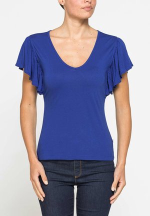 Donna con indosso una maglietta blu reale a scollo a V con maniche corte arricciate e jeans blu scuro, mostrata da metà petto fino ai fianchi su sfondo bianco.