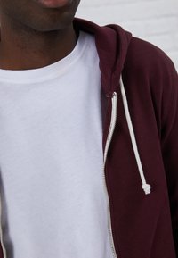 Sudadera burdeos con cremallera y cordones blancos, llevada sobre una camiseta blanca sencilla. La tela parece suave, con una textura lisa y un detallado mínimo.