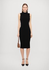 Rochie midi tricotată neagră, cu guler înalt, design fără mâneci, siluetă ajustată și textură netedă. Însoțită de o poșetă mică maro.