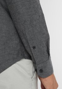 Chemise grise texturée avec un poignet boutonné, comprenant un ourlet incurvé et des boutons noirs. Conçue avec un motif tissé subtil.
