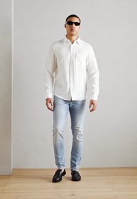 Weißes Langarmhemd mit strukturiertem Finish, hellblaue Jeans und schwarze Schuhe. Das Modell trägt schwarze Sonnenbrillen und posiert vor einem schlichten Hintergrund.