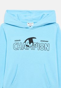 Ανοιχτό μπλε φούτερ με κουκούλα και τσέπη μπροστά, με μαύρο λογότυπο "CHAMPION" και κείμενο που γράφει "Authentic Athleticwear." Μαλακό βαμβακερό υλικό.