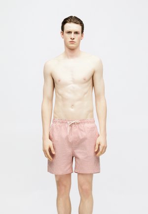 Jeune homme à la peau claire et aux cheveux mouillés coiffés en arrière, portant un short de bain rayé rose et blanc, debout devant un fond blanc uni.