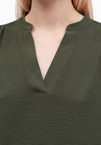 Femme portant un haut vert olive texturé avec un col rond fendu et sans attaches visibles.
