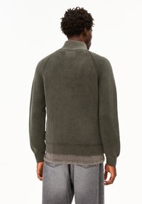 Mann mit kurzen lockigen Haaren, der einen olivgrünen gerippten Pullover und verwaschene graue Jeans trägt, steht mit dem Rücken zu einem weißen Hintergrund.