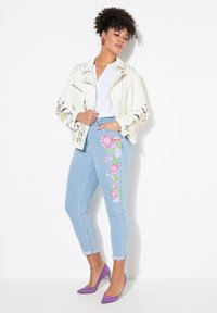 Bílá vyšívaná bunda s nýty, modré džíny s vysokým pasem a květinovým vzorem a fialové podpatky. Oblečení má neformální, vrstvený vzhled.